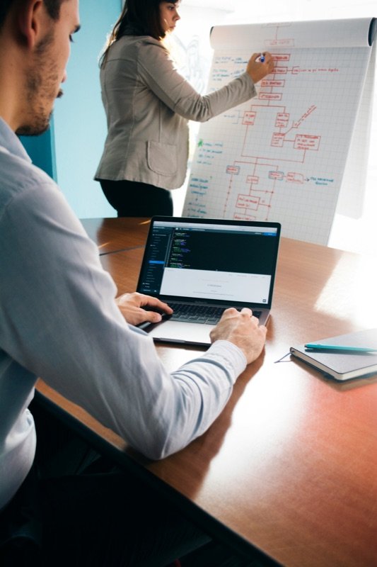 Developer coding on a laptop while a colleague maps a flowchart on a whiteboard